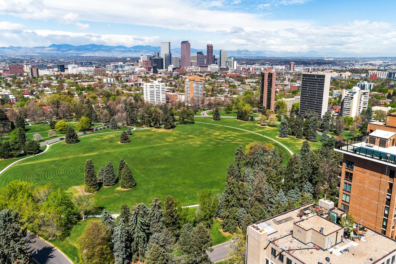 Aerial view of Cheesman Park in Denver, CO featuring expansive green lawns, walking paths, surrounding residential buildings, and the downtown skyline with mountain views in the distance.