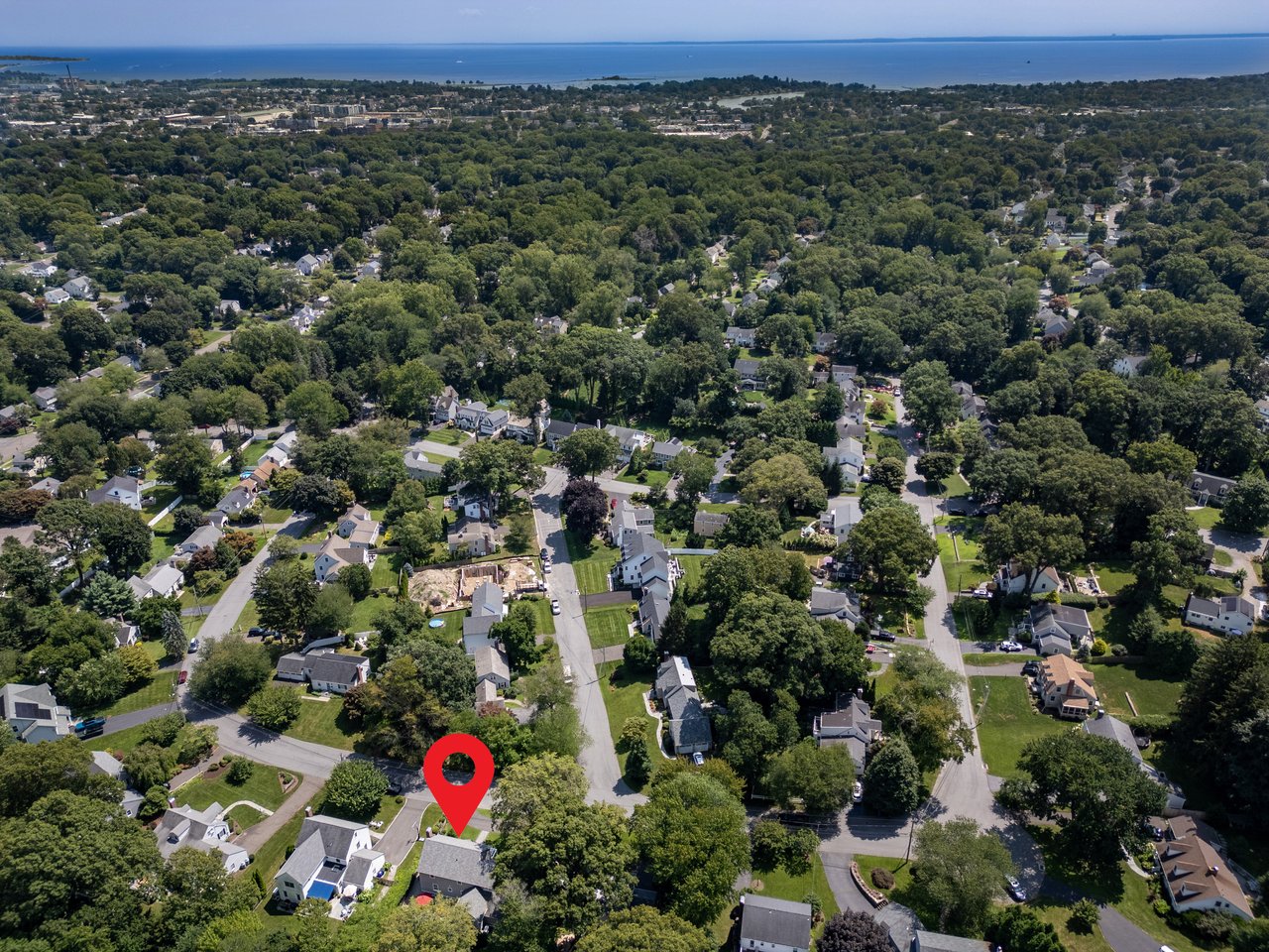 Aerial view of a tree-lined neighborhood, with a red marker above one home and water on the horizon.