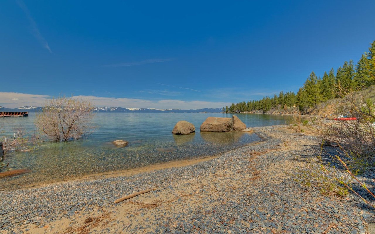 Lake Tahoe shoreline with clear water, boulders, and snow-dusted mountains beyond.