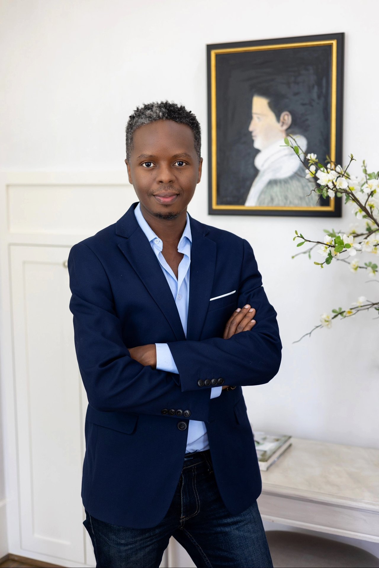 Stephen Mweka standing with arms crossed in a navy blazer beside a framed portrait and flowering branches.