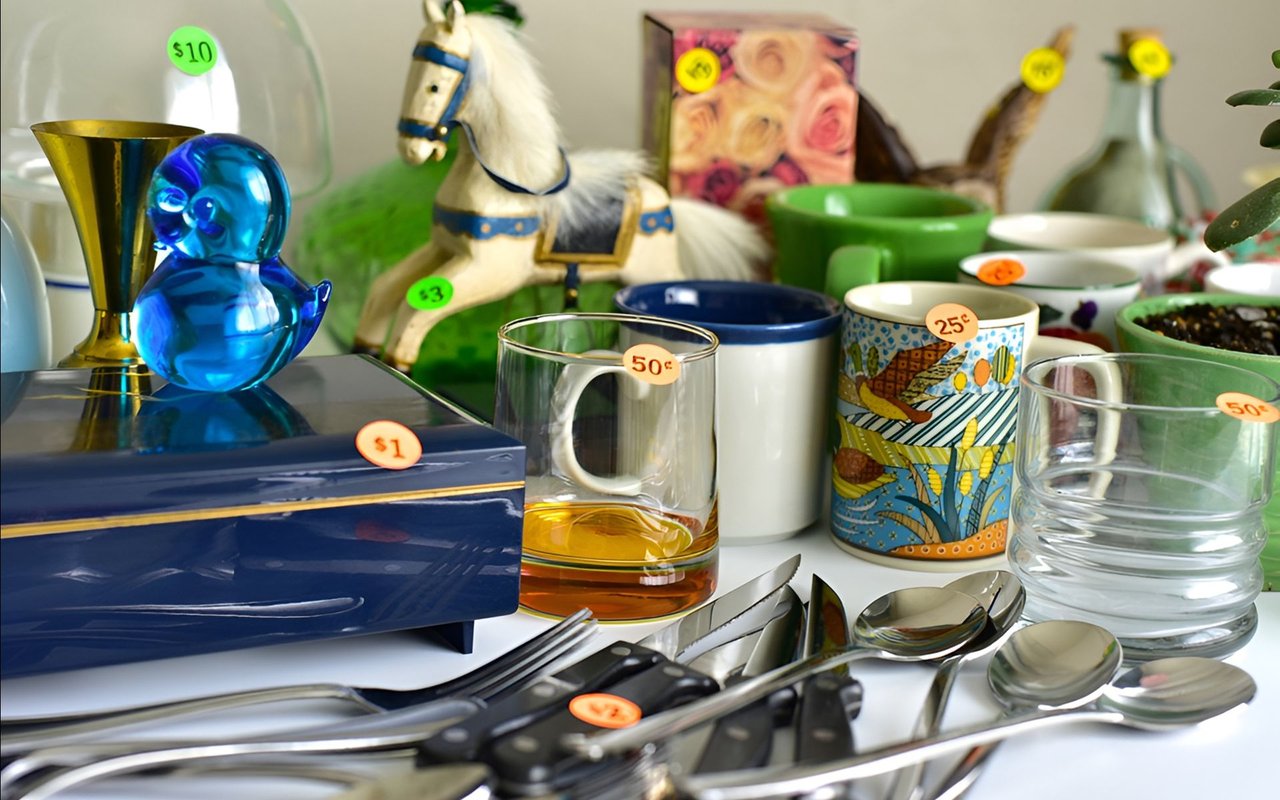 Yard-sale table crowded with priced glassware, bowls, cutlery, a toy horse, and a blue glass figurine.