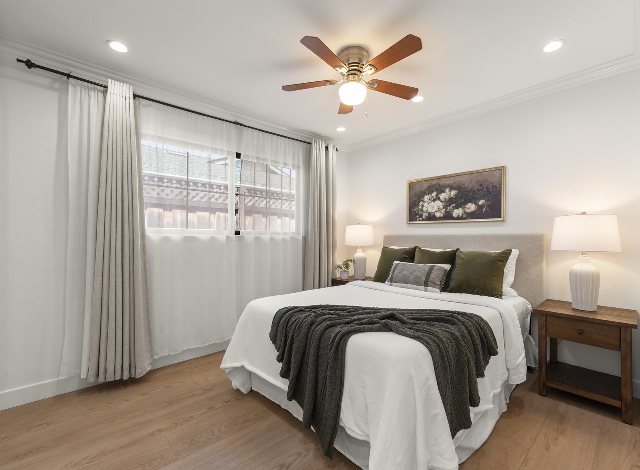 Staged bedroom with a white bed, dark throw, wooden ceiling fan, and large curtained window.