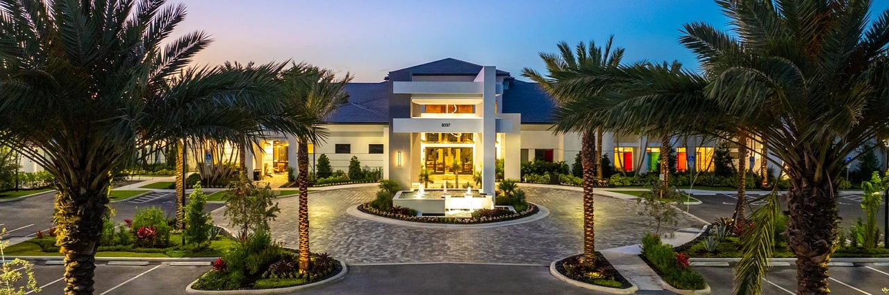 Modern clubhouse entrance with a circular driveway and palm trees at dusk.