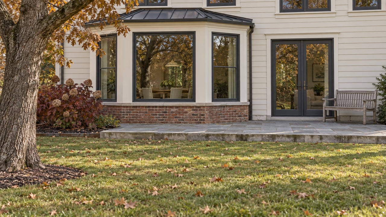 Autumn lawn leading to a limestone terrace and glazed breakfast bay on an ivory-sided home, with a mature bur oak and burgundy oakleaf hydrangea.