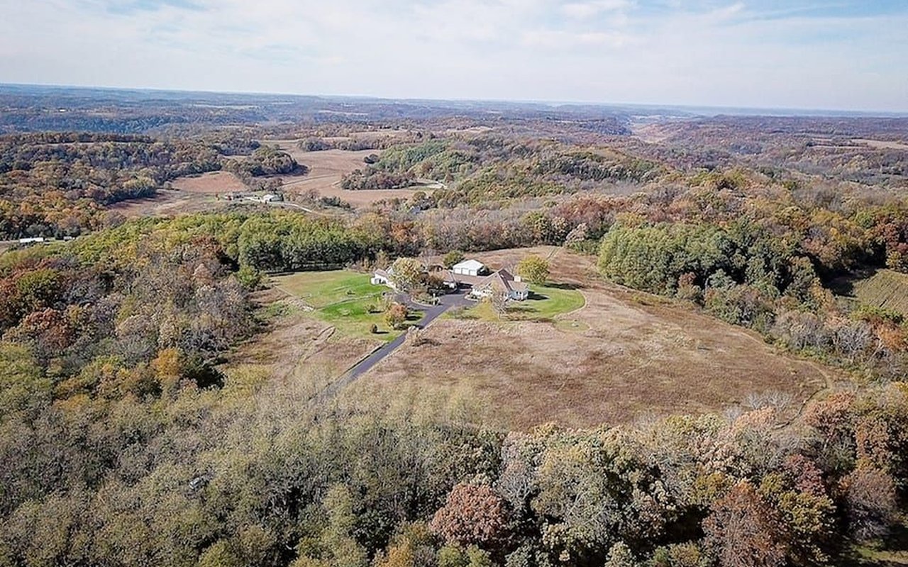 Rural home and outbuildings amid open fields and autumn-colored wooded hills.
