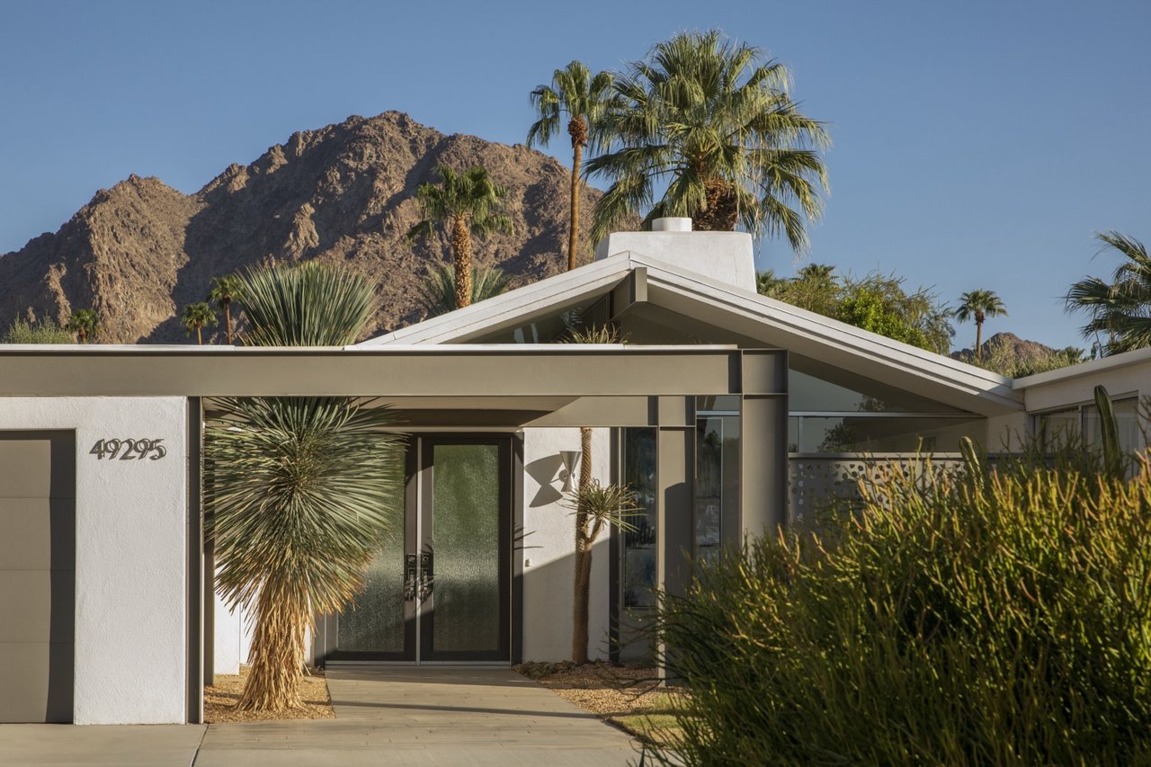 Modernist home with a broad gabled roof, desert landscaping, palm trees, and a mountain backdrop.