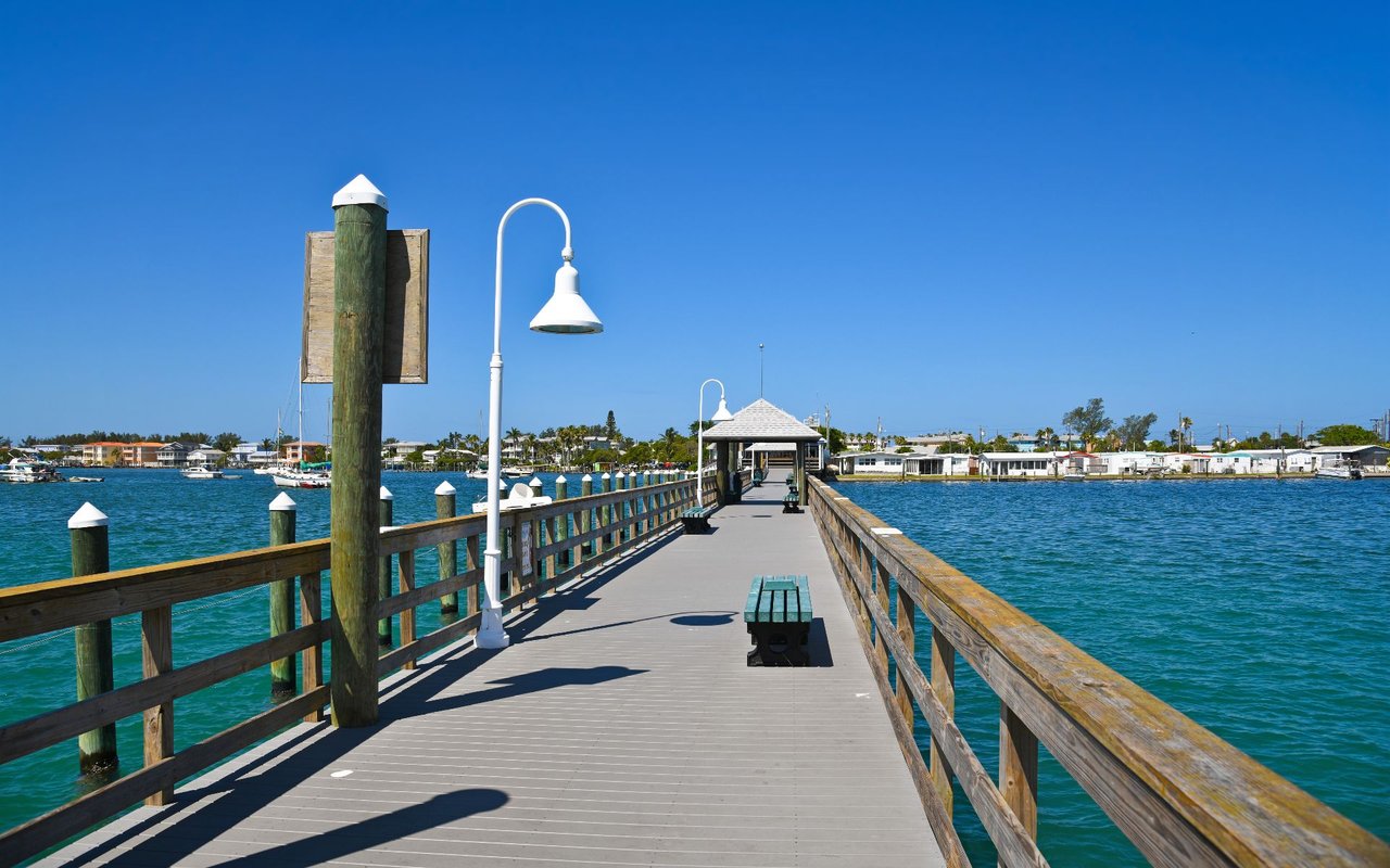 Wooden pier extends over blue water toward a covered pavilion, with homes along the shore.