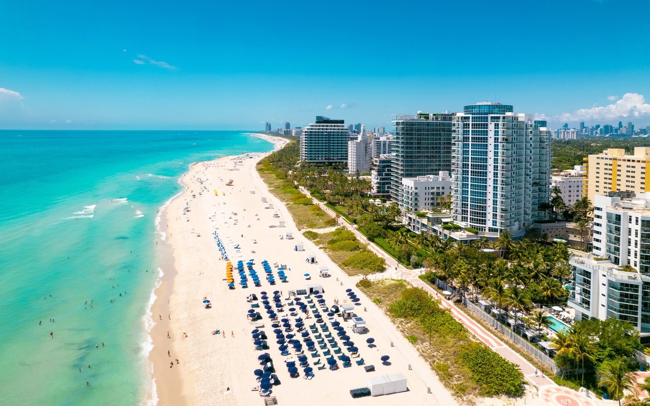 Aerial view of beachfront condominiums beside turquoise water and a sandy beach lined with umbrellas.