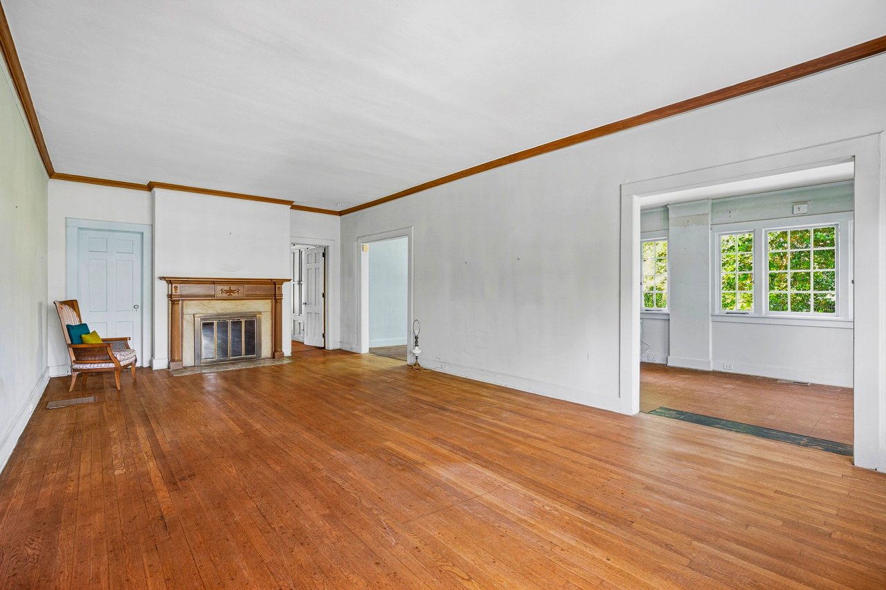 Living room with hardwood floors, a wood-trimmed fireplace, and an adjoining room with tall windows.