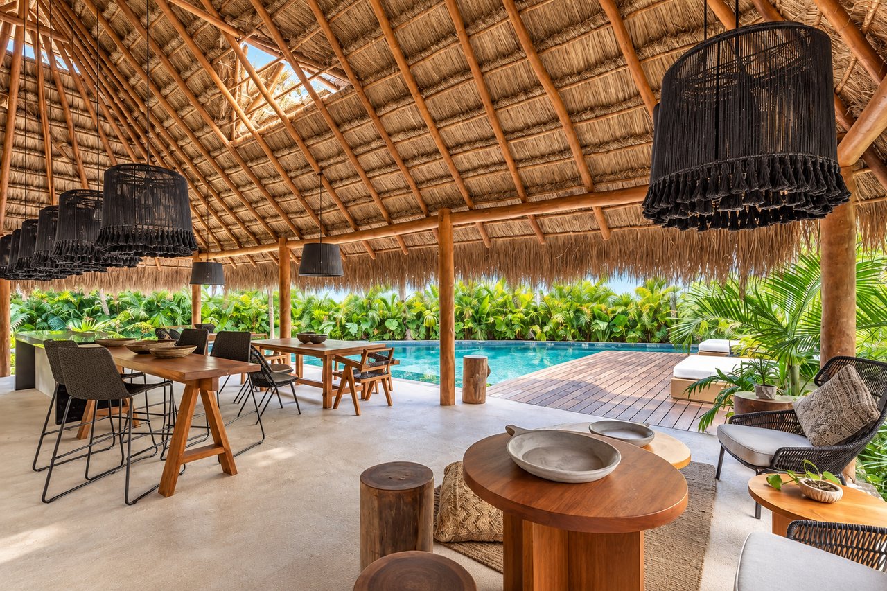 Covered outdoor dining and lounge area beside a swimming pool under a thatched roof.