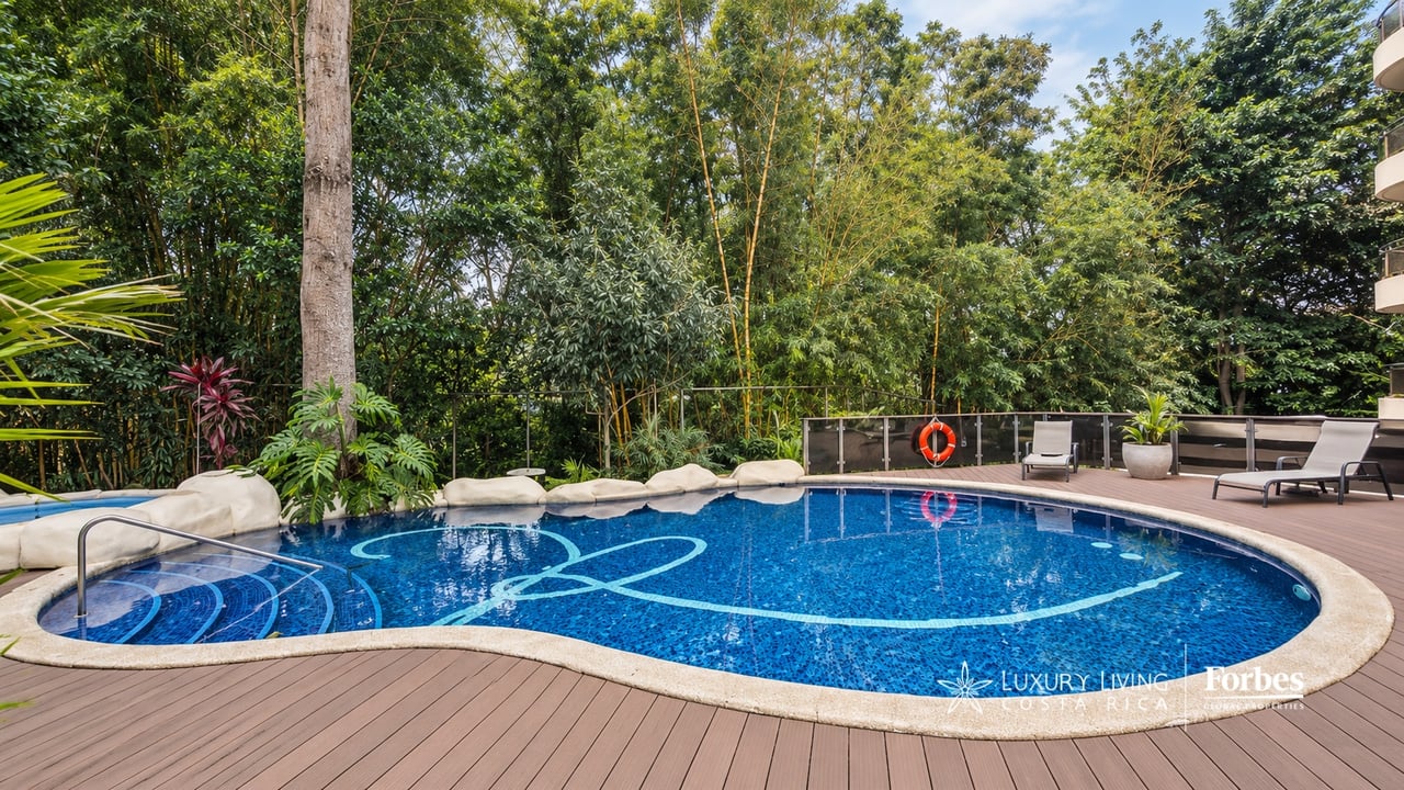 Outdoor pool with curved entry steps, a wooden deck, lounge chairs, and dense tropical greenery.