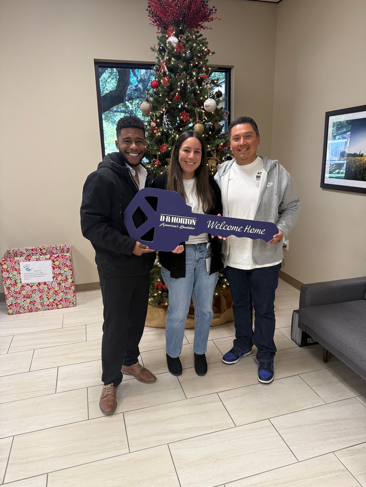 Three adults celebrate with an oversized Welcome Home key in front of a decorated Christmas tree.