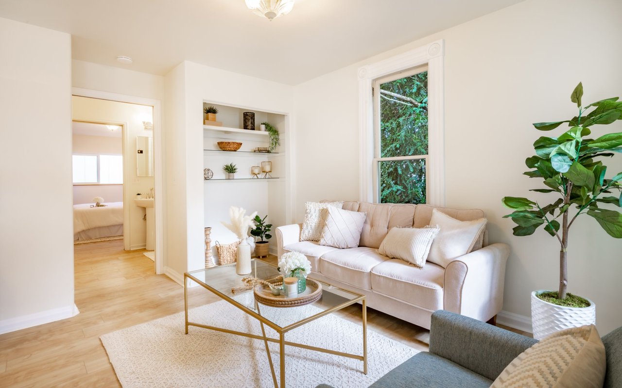 Staged living room with a cream sofa, glass coffee table, built-in shelves, and a window overlooking greenery.