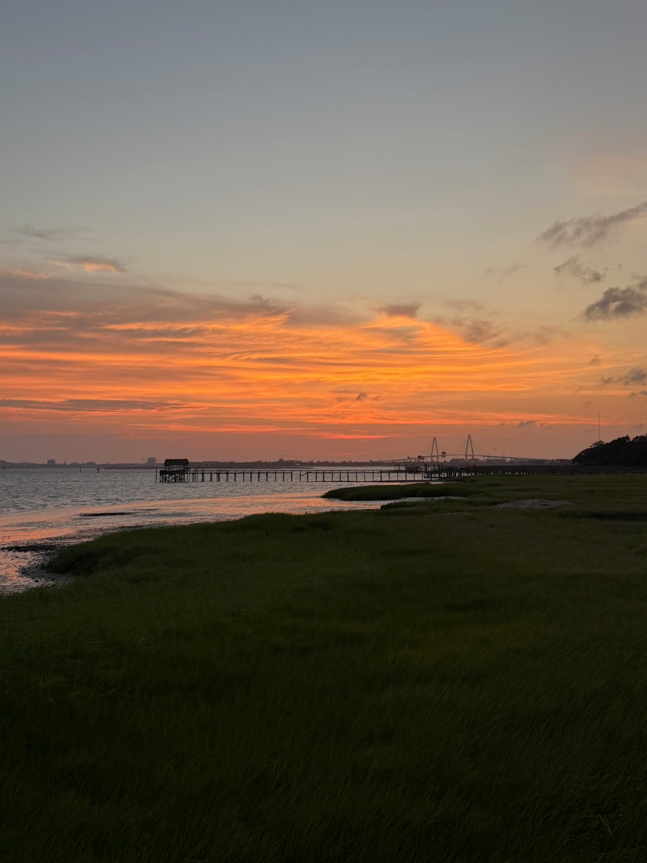 Orange sunset over a coastal marsh, calm water, and a long pier, with a bridge on the horizon.
