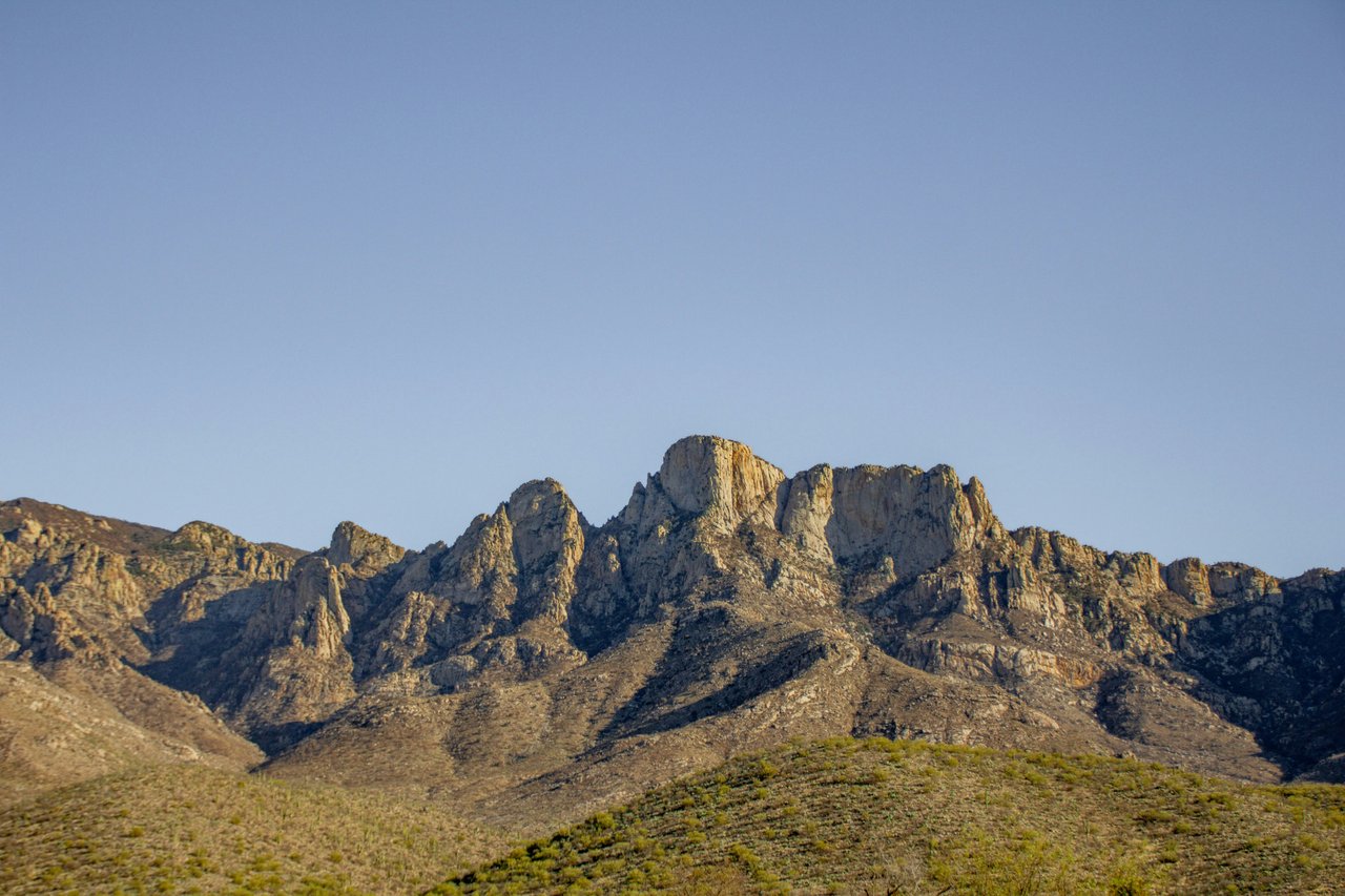 Rugged, sunlit mountain peaks rise above a dry, scrub-covered foothill beneath a clear blue sky.