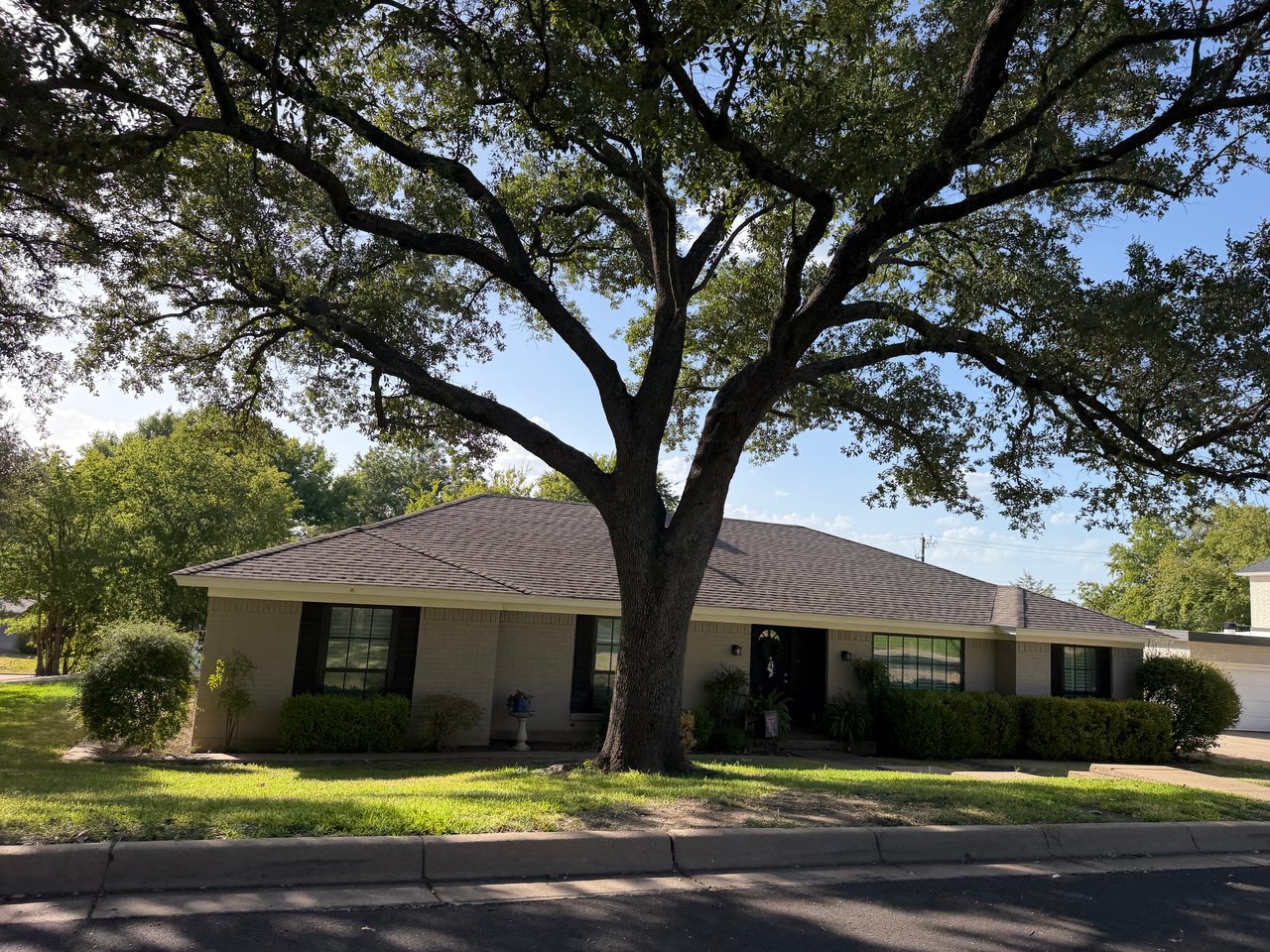 Single-story brick home shaded by a large mature tree, with a low hipped roof and front shrubs.