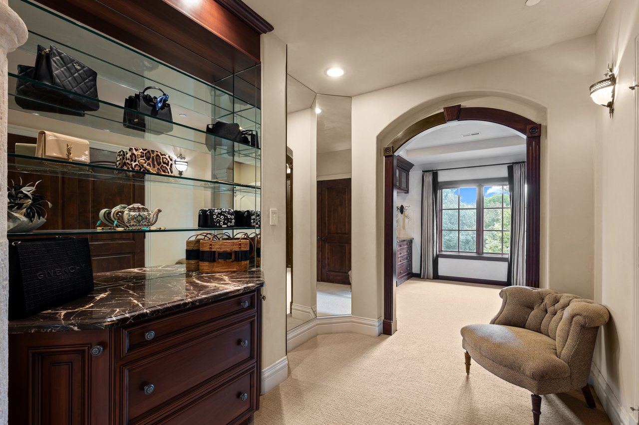 Dressing room with glass display shelves, dark wood cabinetry, marble counter, and a tufted chair.