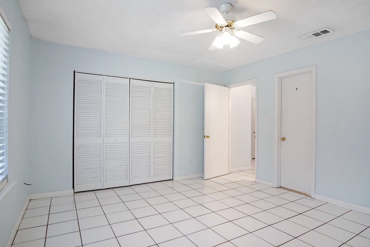 A bright, empty room with light blue walls, white tiled floor, and a ceiling fan. Closed white closet doors and an open white door are visible. Peaceful ambiance.