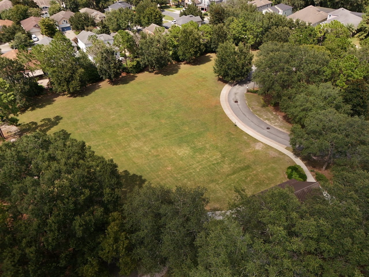 Aerial view of a grassy neighborhood green bordered by trees, homes, and a curved road.