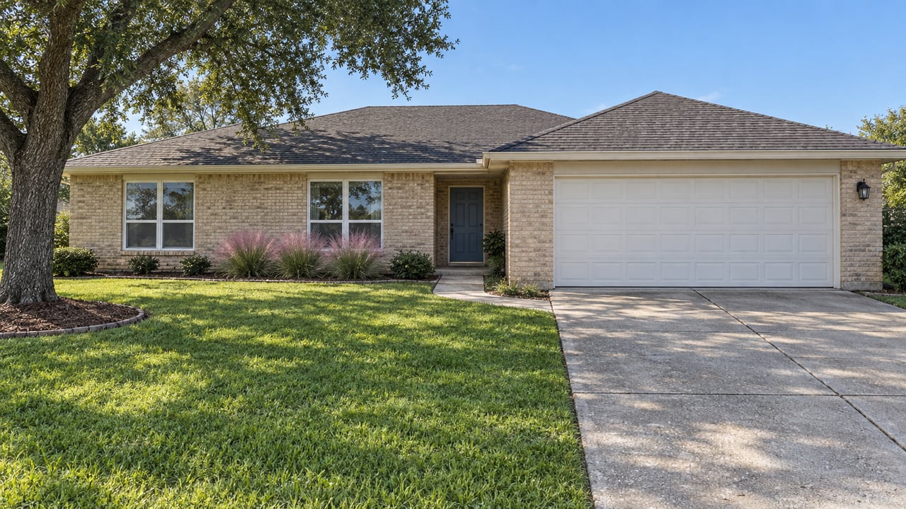 Single-story buff-brick home with a blue recessed entry, white two-car garage, deep green lawn, and a mature live oak in bright daylight.