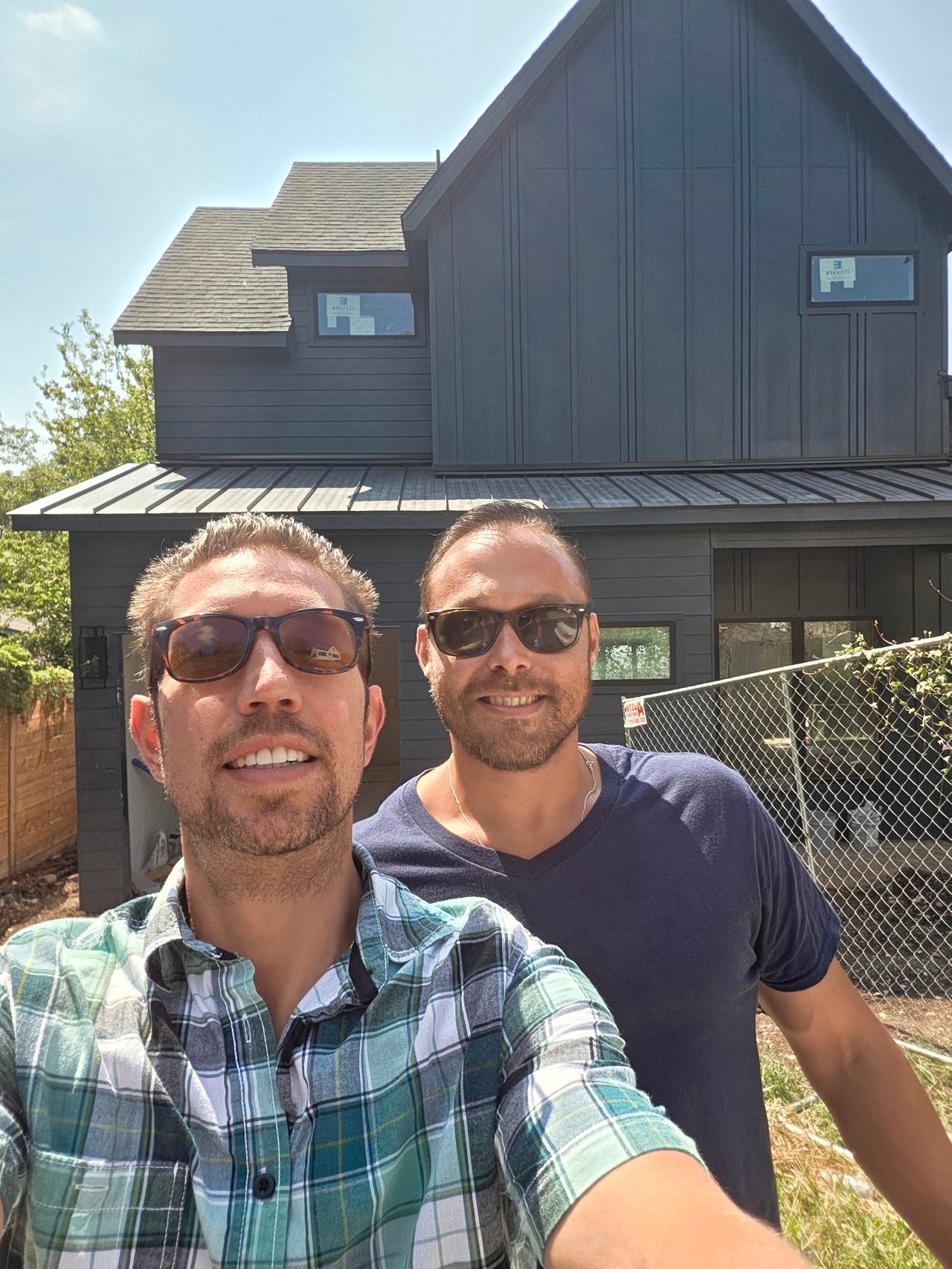 Two men in sunglasses take a selfie in front of a dark gray modern house.