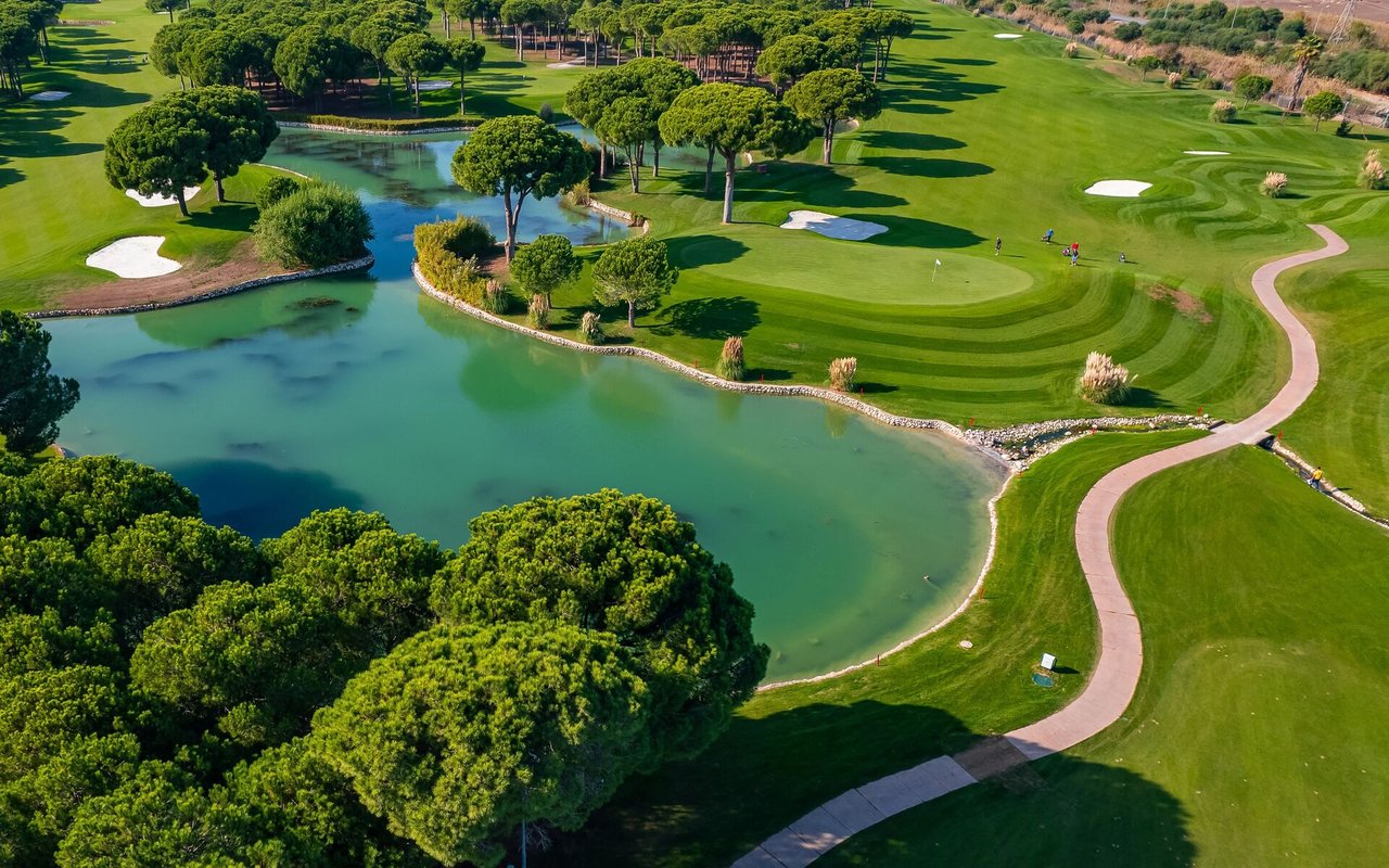 Aerial golf course with a pond, tree-lined fairways, sand bunkers, and a winding cart path.