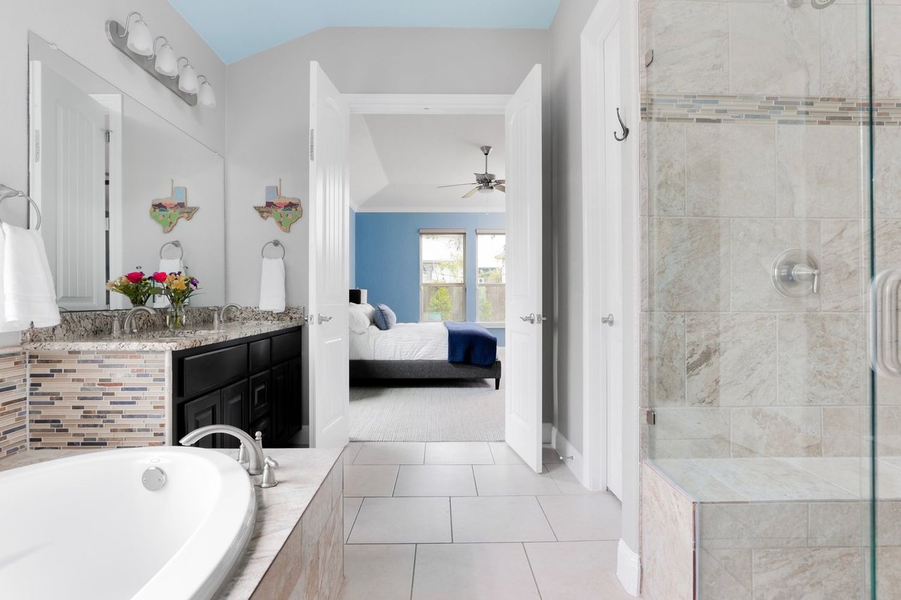 Primary bathroom with soaking tub, dark double vanity, tiled shower, and bedroom visible through open doors.