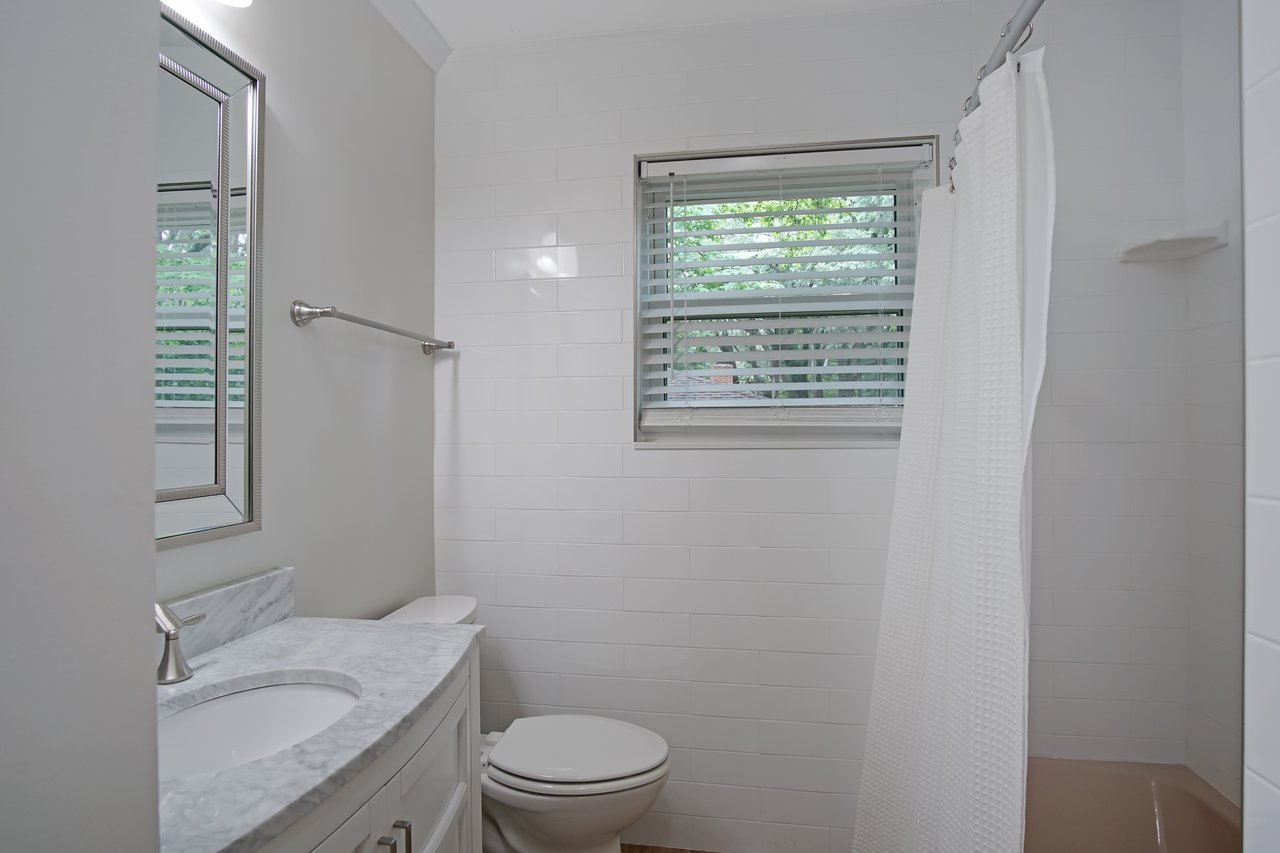 Small, bright bathroom with a white marble-topped vanity, oval mirror, and chrome faucet. Features a toilet, window with blinds, and shower with a white curtain. Quiet ambiance.