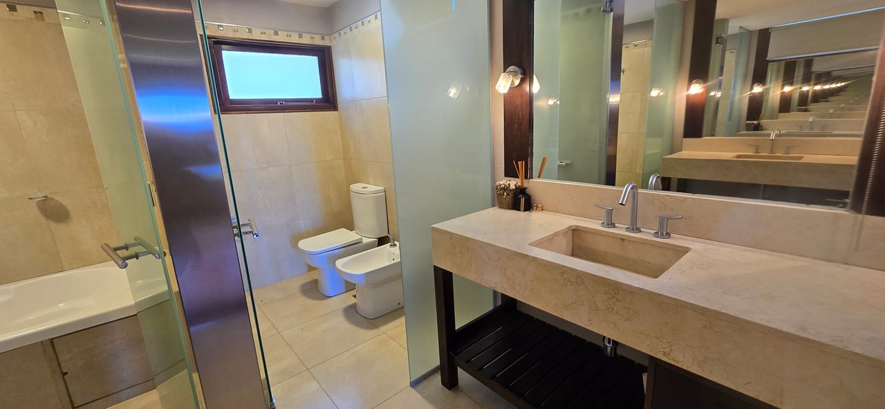 Modern bathroom with a beige stone double-sink vanity, glass shower, bathtub, toilet and bidet.