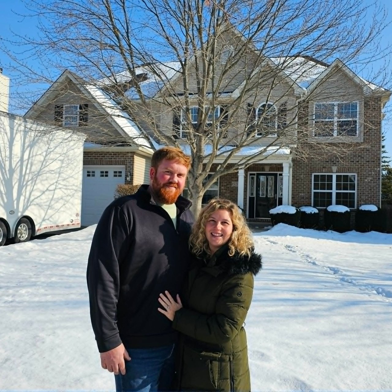 Couple standing in a snowy driveway in front of a large brick house.
