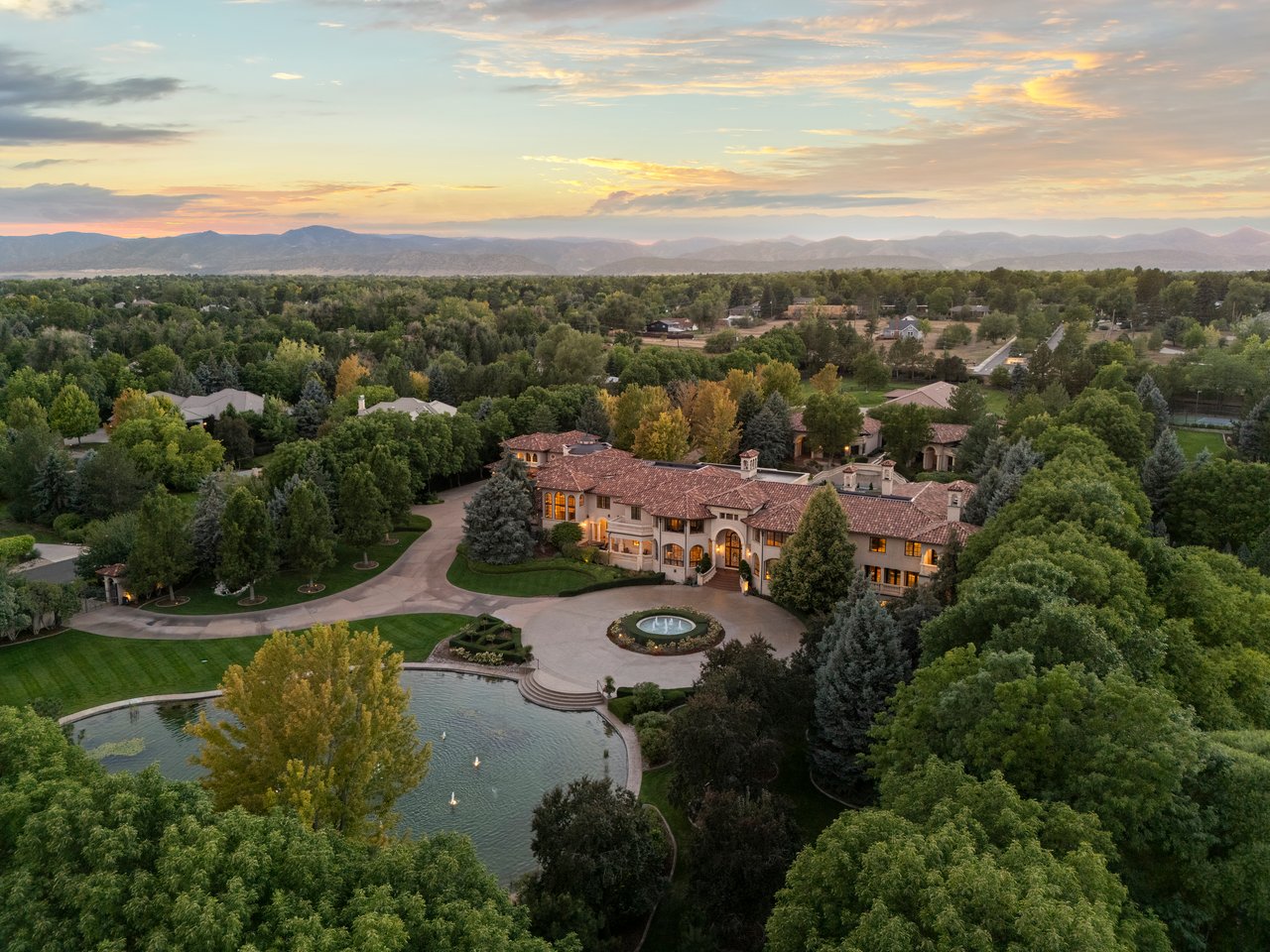 Aerial view of a large mansion beside a pond, with landscaped grounds and distant mountains.