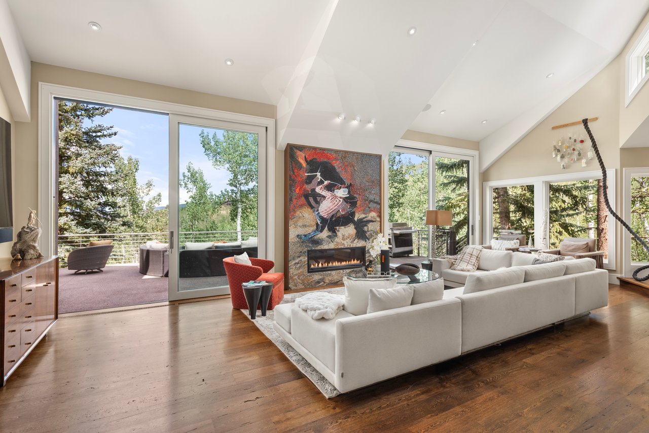 Living room with a white sectional, colorful fireplace artwork, and windows opening to a tree-lined terrace.