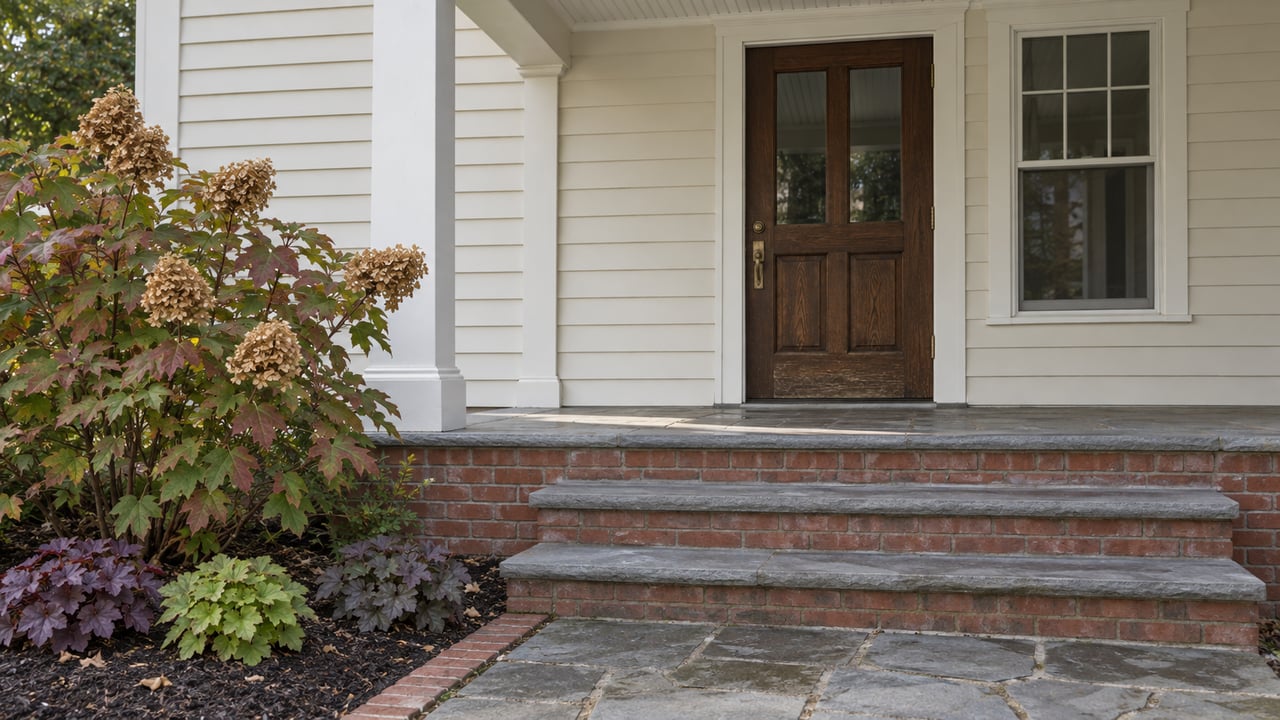 Traditional home's covered entry with cream lap siding, oak door, brick and stone steps, and oakleaf hydrangea beside the walk.
