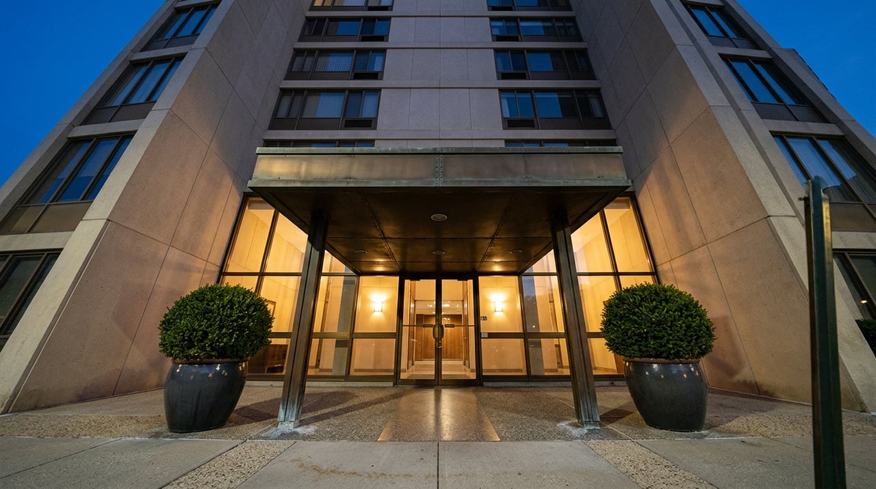 Postwar tower entrance with a bronze canopy, glowing lobby windows, and clipped shrubs in matching planters.