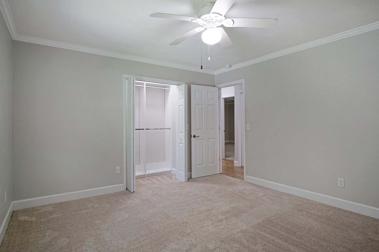 Empty room with beige carpet and light gray walls. Open closet doors and hallway entrance are visible. Ceiling fan with light is turned on, creating a calm atmosphere.