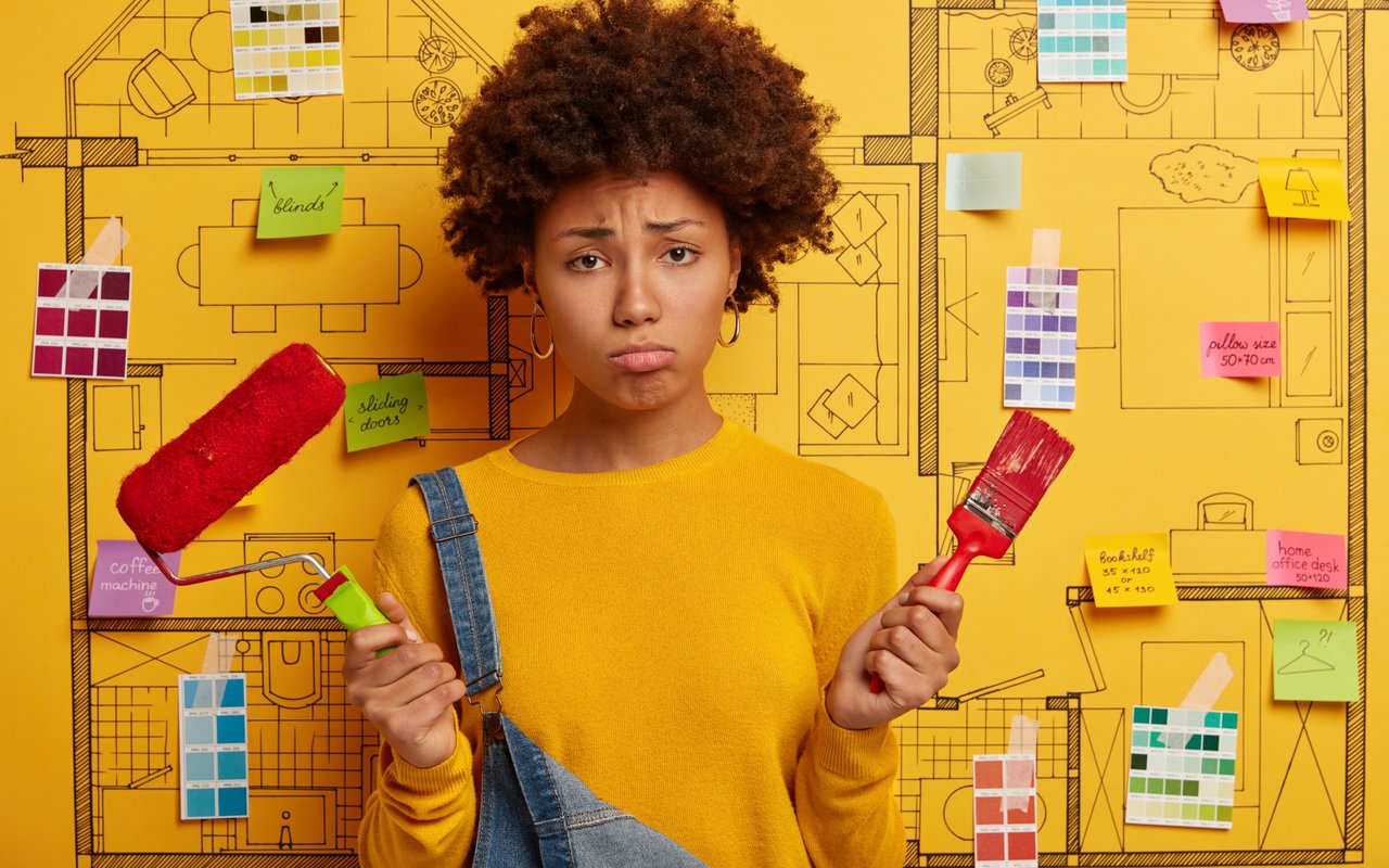 Worried woman holding a paint roller and brush in front of renovation plans and color swatches.