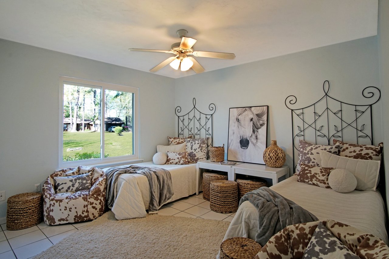 Cozy bedroom with two beds and brown cow-print accents. A large window shows a green outdoor view. Rustic decor includes wicker baskets and a horse photo.