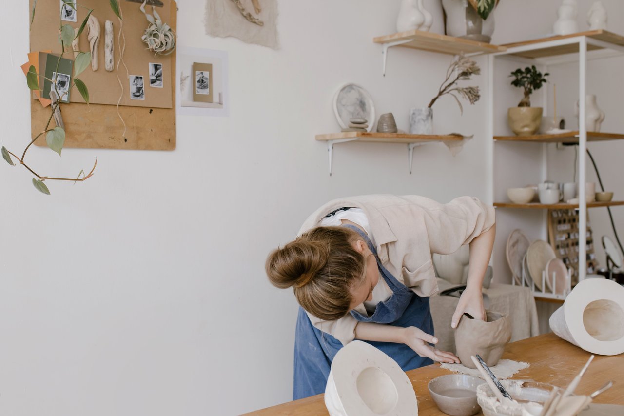 A woman shapes a clay vessel at a pottery studio worktable.