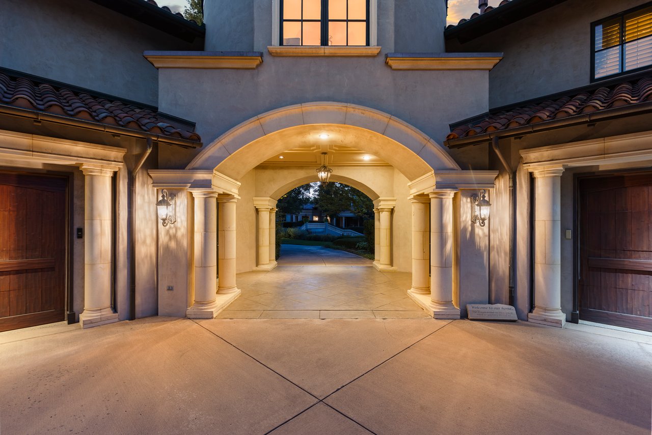 Luxury home entrance with an illuminated stone arch, columns, and tiled roofs at dusk.