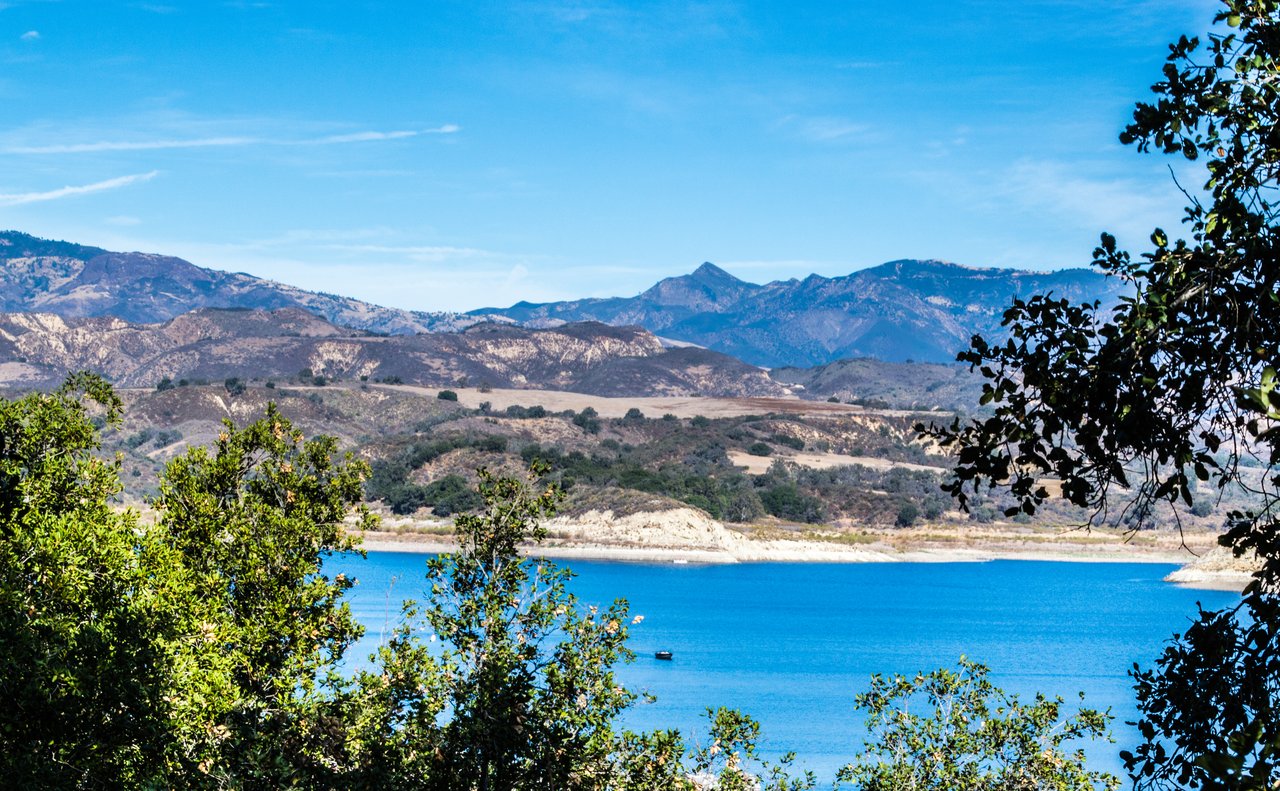 Blue lake bordered by dry hills, with rugged mountains in the distance and leafy branches in the foreground.