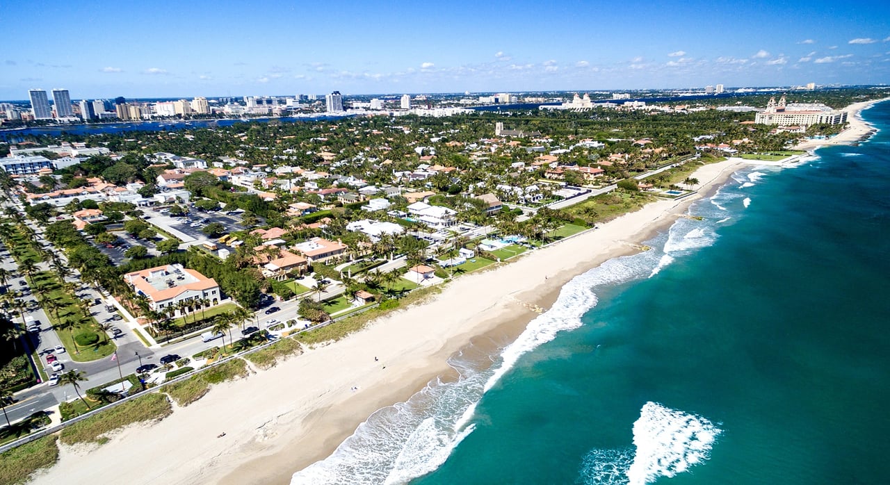 Aerial view of Palm Beach island’s sandy Atlantic beach, turquoise water, palm-lined homes, and distant skyline.