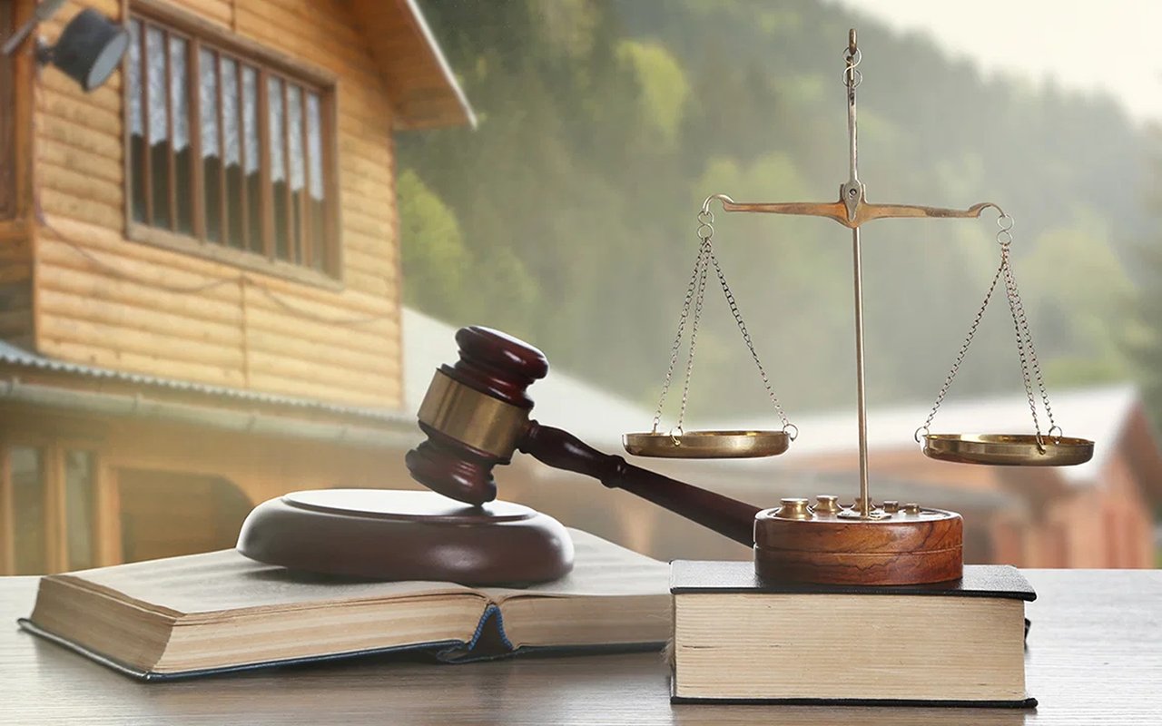 Gavel and balance scales resting on books, with a wooden house in the background.