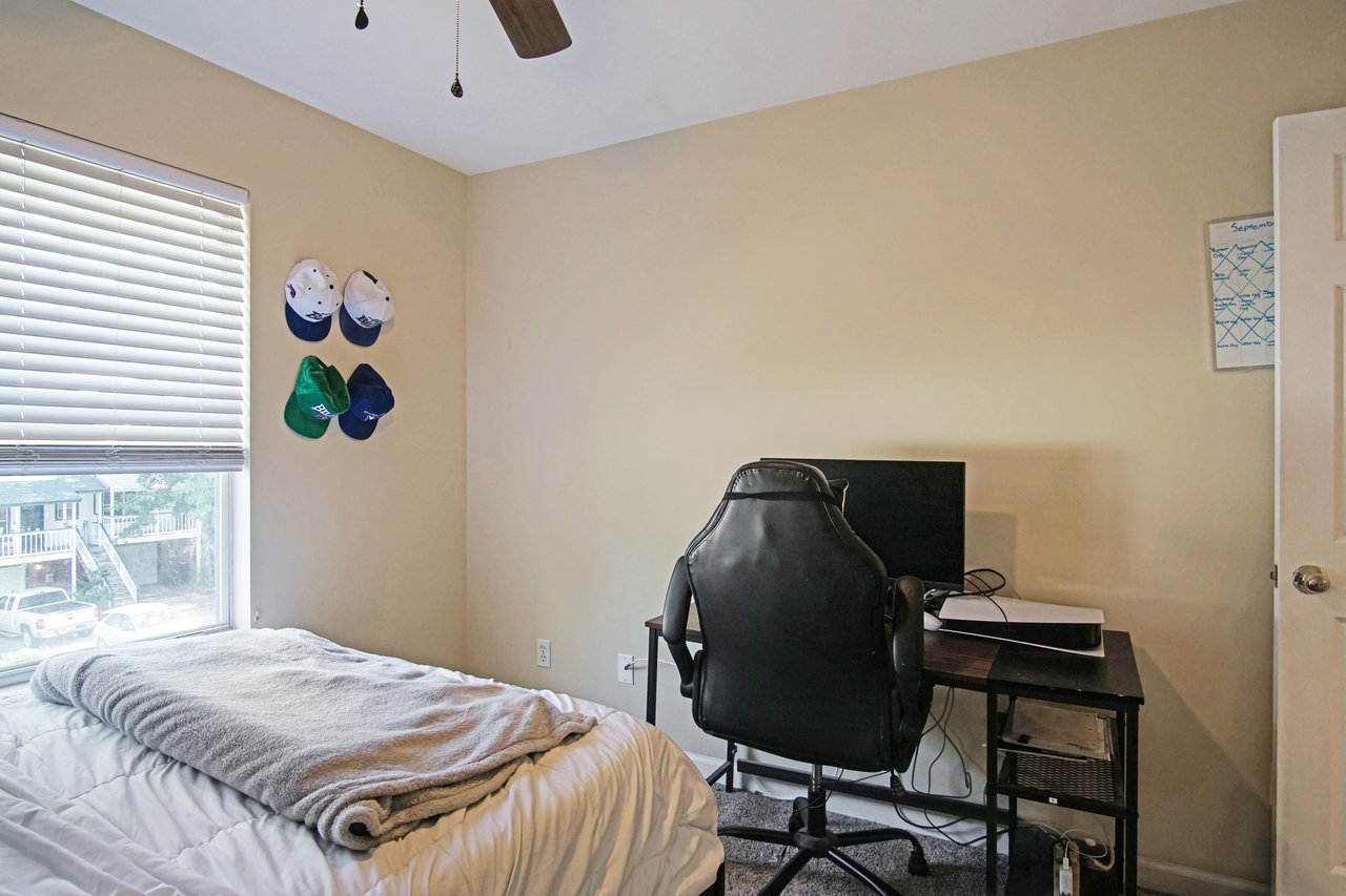 Bedroom at 529 Collinsford Road, Tallahassee, Florida, featuring a single bed with white bedding, a desk setup with a computer and office chair, and wall-mounted baseball caps. The room has beige walls, carpet flooring, and a large window with blinds letting in natural light.