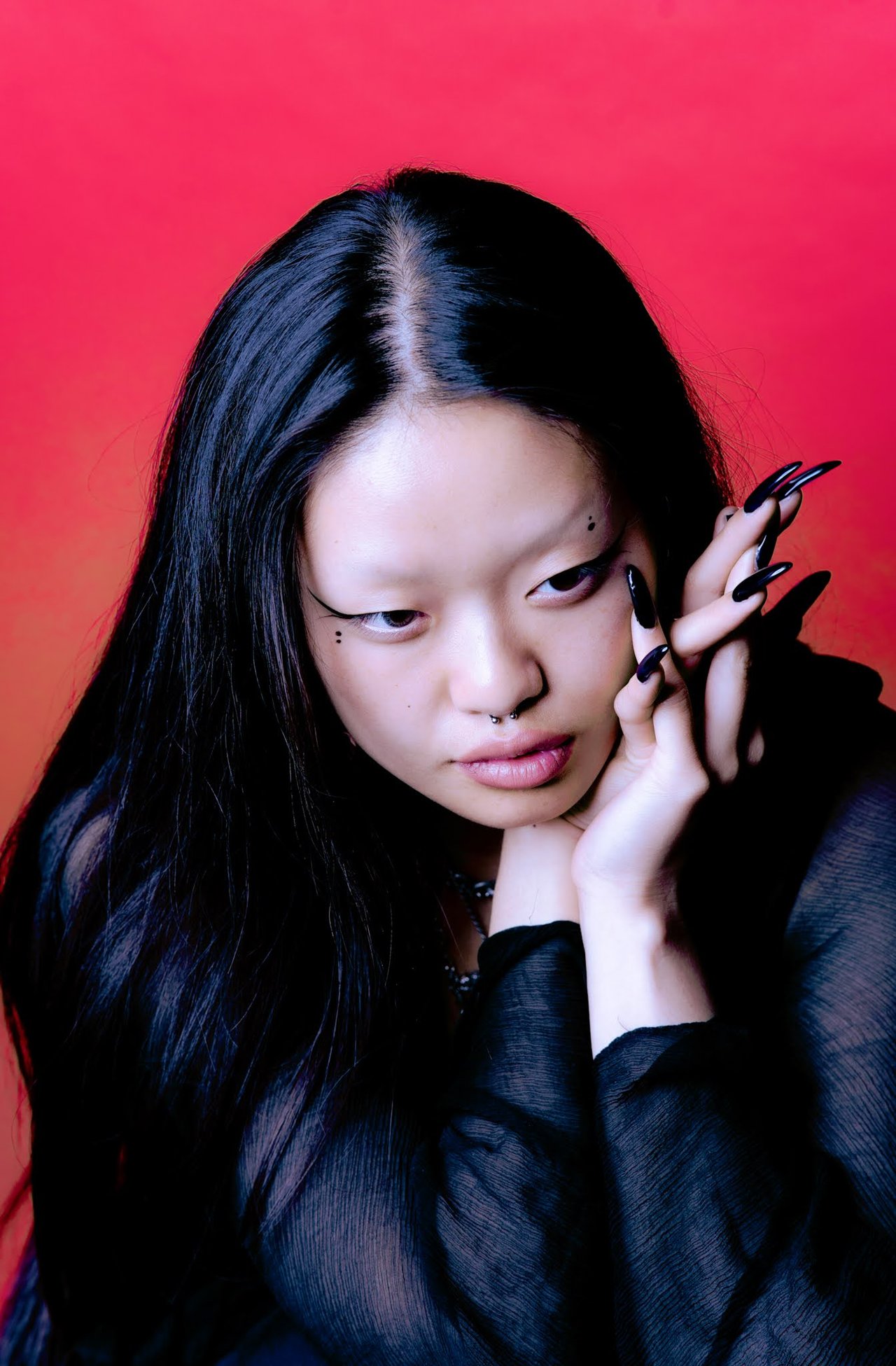 Dancer Jin poses against a vivid red backdrop, framing her face with long black nails.