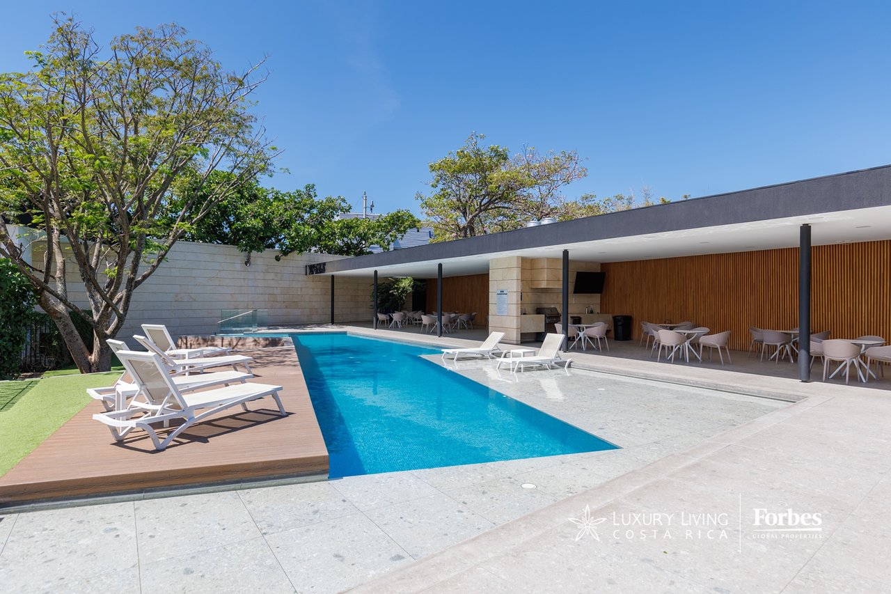 Contemporary home’s long swimming pool beside a covered outdoor dining area and wood deck.