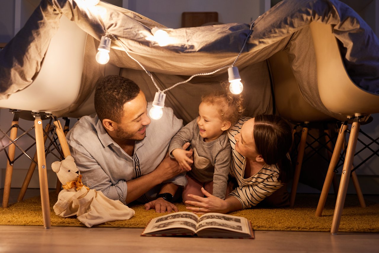 A family of three enjoys time under a blanket fort, immersed in a book with string lights overhead, much like finding solace with real estate agents guiding new beginnings.