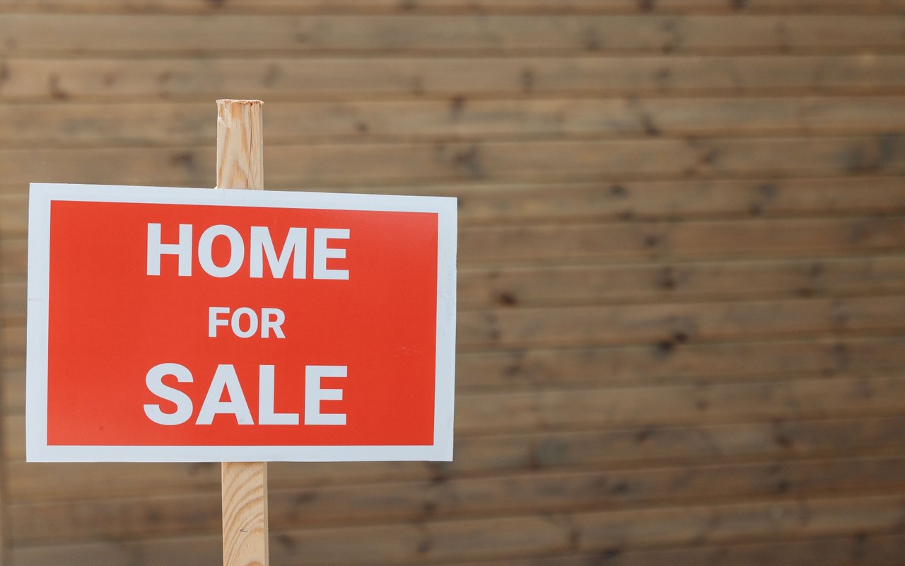Red-and-white HOME FOR SALE sign against a wooden plank wall.