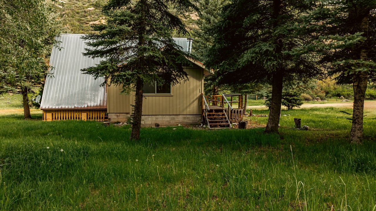 Tan cabin with a metal roof and wooden steps in a grassy, tree-shaded clearing.