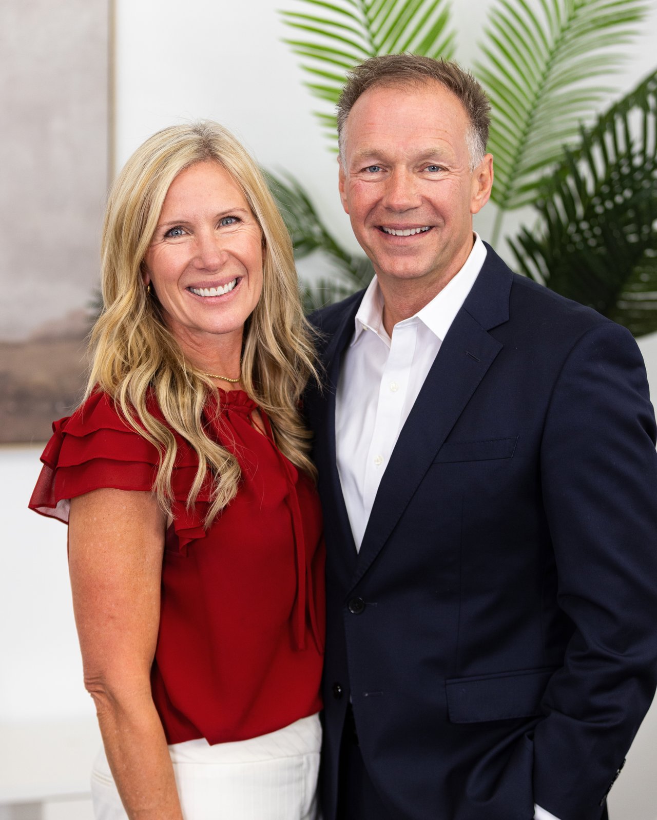 A man in a navy suit and a woman in a red blouse pose together in front of palm leaves.