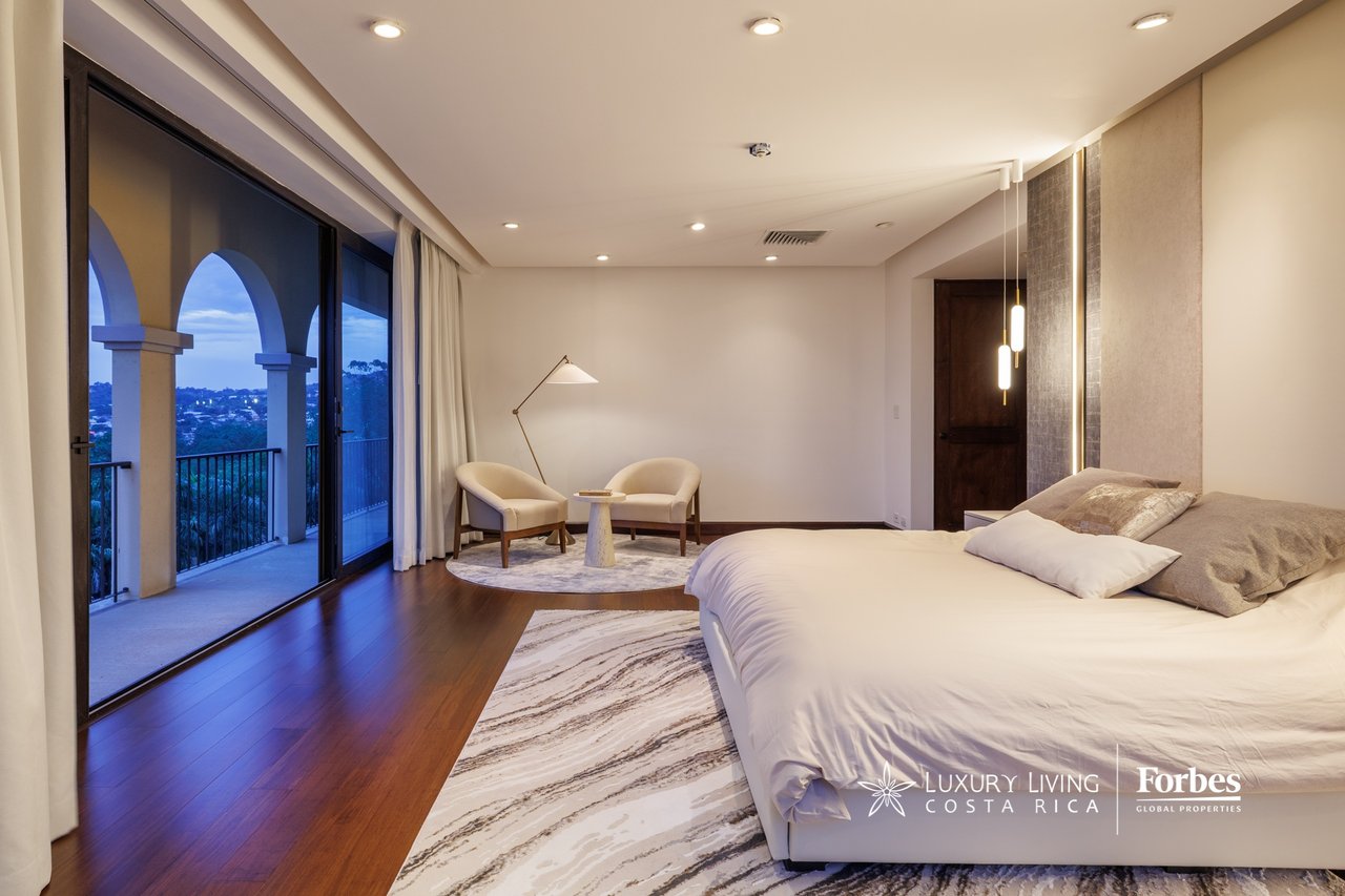 Penthouse bedroom with a large bed and arched balcony doors overlooking Escazú at dusk.
